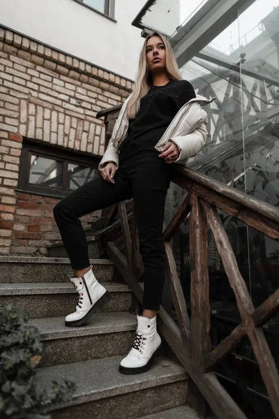 Modelo elegante de uma jovem mulher em preto-branco outerwear elegante em couro outono botas da moda fica em uma escadaria vintage. Menina bonito está descansando na cidade perto do corrimão de madeira. Estilo casual — Fotografia de Stock