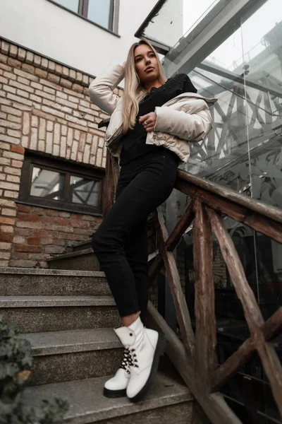 Αστικό μοντέλο μόδας μιας χαριτωμένης νεαρής γυναίκας με ανοιξιάτικα-φθινοπωρινά ρούχα σε μοντέρνες μπότες που ποζάρουν στην πόλη σε μια σκάλα με παλιά ξύλινα κάγκελα. Ελκυστική κοπέλα σε ένα κομψό εξωτερικά ενδύματα στέκεται σε εξωτερικούς χώρους. — Φωτογραφία Αρχείου