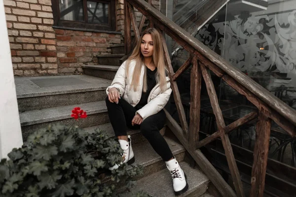 Modèle jeune femme mignonne à la mode dans une veste blanche élégante en jean noir en cuir bottes blanches à la mode se trouve sur un vieil escalier en pierre dans la ville. Attrayant fille moderne en plein air dans un jour d'automne . — Photo