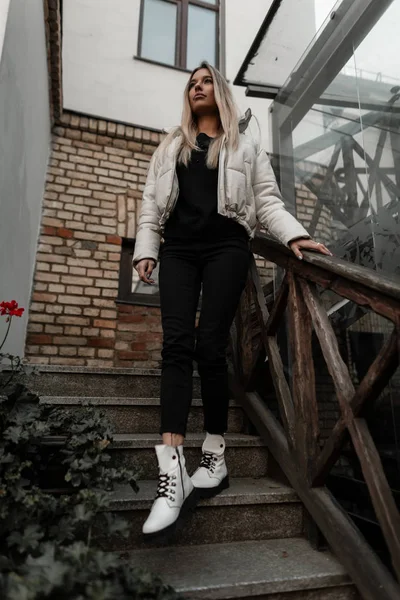 Όμορφη νεαρή γυναίκα με μακριά ξανθά μαλλιά σε ένα μοντέρνο λευκό σακάκι σε μαύρα ρούχα σε δερμάτινες κομψές φθινοπωρινές μπότες ποζάρουν σε μια vintage σκάλα στην πόλη. Χαριτωμένο κορίτσι σε μοντέρνα εξωτερικά ενδύματα — Φωτογραφία Αρχείου