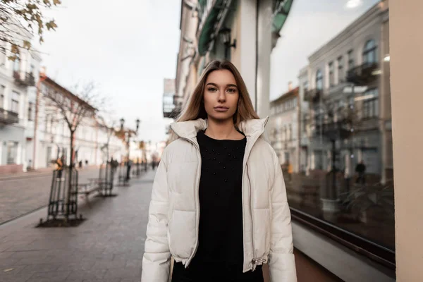 Modelo bastante europeo de mujer joven en una chaqueta blanca de moda en vaqueros negros con estilo en un suéter de punto se encuentra cerca de un edificio vintage. Atractiva chica rubia moderna disfruta de un paseo por la ciudad . — Foto de Stock