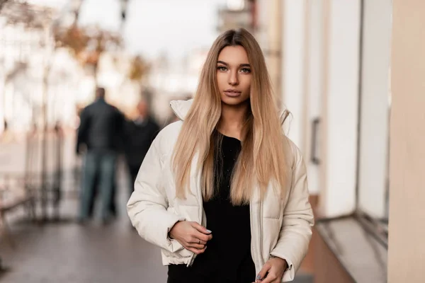 Mulher jovem loira glamourosa com lábios sexy em um suéter elegante preto em uma jaqueta de inverno branca na moda anda ao ar livre perto de um edifício vintage. Bonito modelo menina fica na rua. Moda jovem . — Fotografia de Stock