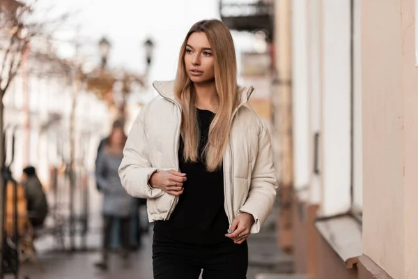 Joven rubia glamorosa europea en una chaqueta blanca de moda en vaqueros negros en un suéter se encuentra al aire libre cerca de un edificio vintage. Atractiva modelo de chica moderna camina por la calle. Moda . —  Fotos de Stock