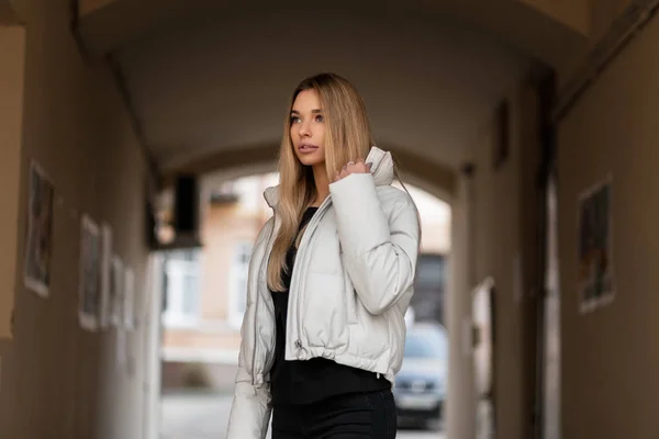 Modello di una bella giovane donna in una giacca bianca alla moda in un maglione nero all'aperto in città vicino a un edificio d'epoca. Urban ragazza elegante cammina per strada in abiti alla moda. Stile primaverile . — Foto Stock