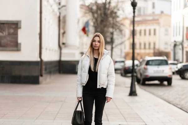 Giovane donna alla moda bionda in elegante capispalla bianco-nero con una borsa di moda in pelle sta camminando per strada in una calda giornata autunnale. Carino modello ragazza alla moda in abiti giovanili all'aperto. Look casual . — Foto Stock