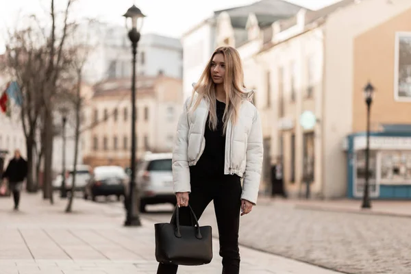 Modelo de moda moderna de una mujer joven en una chaqueta blanca de moda en jeans con estilo con una bolsa de cuero de moda se encuentra en la calle en un día de otoño. Chica rubia con estilo en una ropa de juventud en la ciudad — Foto de Stock