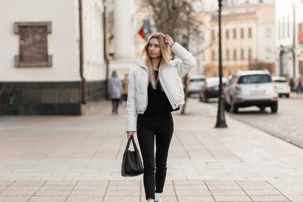 Ładna miejska młoda kobieta w modnej białej kurtce w klasycznych czarnych dżinsach z czarną stylową skórzaną torbą na ulicy. Atrakcyjny model dziewczyny mody podróżuje po mieście. Jesienny styl. — Zdjęcie stockowe