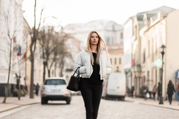 Modelo de mulher jovem urbano magro com cabelo loiro longo em uma jaqueta branca na moda em calças de ganga pretas elegantes em uma bolsa de couro na moda caminha ao longo de uma rua da cidade em um dia de outono quente. Menina moderna está andando . — Fotografia de Stock