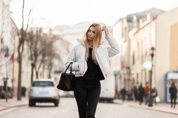 Mulher loira jovem atraente está andando na cidade em uma jaqueta branca elegante em jeans pretos vintage com uma bolsa de couro. Bonita modelo menina caminha pela rua em um dia quente de primavera . — Fotografia de Stock