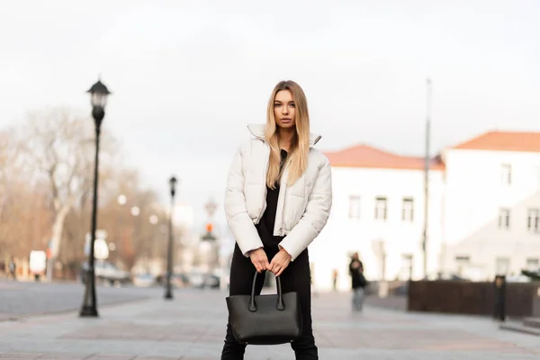 Elegante modello di moda di una giovane donna in una giacca bianca alla moda in jeans alla moda con una borsa alla moda in pelle si erge per strada in una giornata autunnale. Ragazza bionda carina in abiti da gioventù in città . — Foto Stock