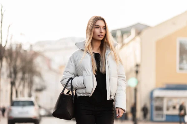 Charmante junge blonde Frau in schwarz-weißer stylischer Kleidung mit lederner Vintage-Tasche läuft an einem Herbsttag die Straße entlang. europäisches Model genießt einen Spaziergang in der Stadt. — Stockfoto