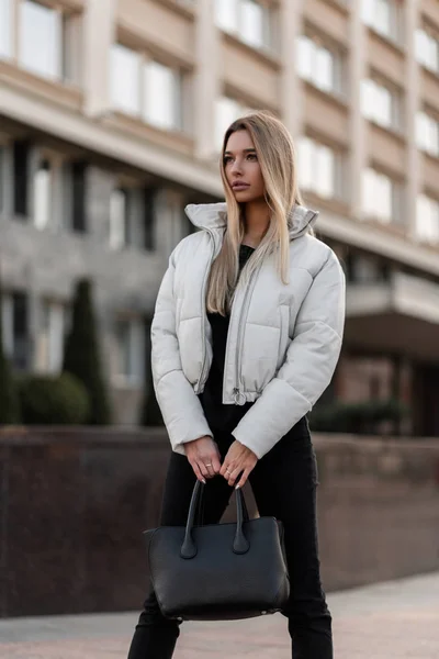 Young model of a beautiful woman with stylish black bag in a fashionable white jacket in jeans stands outdoors on an autumn day near a modern building. Girl in a trendy clothes walks in the city. — Stock Photo, Image