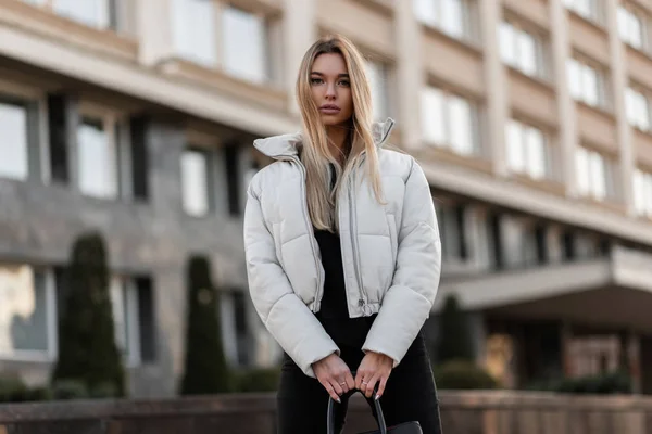 Mulher elegante sexy bonito em uma jaqueta branca elegante fica na cidade no fundo de um edifício moderno. Europeu modelo de moda menina na moda outwear outono-primavera posando na rua . — Fotografia de Stock