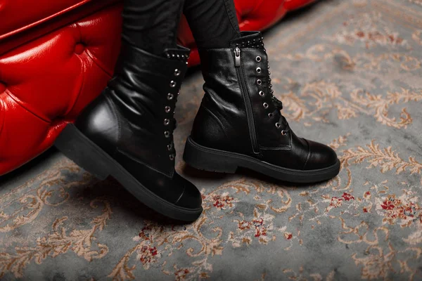 Detailní záběr ženských nohou v módních džínách v sezónních stylových kožených černých krajkových botách v místnosti. Moderní dívka v módních botách sedí na červeném měkkém křesle v obchodě. Dámská zimní obuv. — Stock fotografie