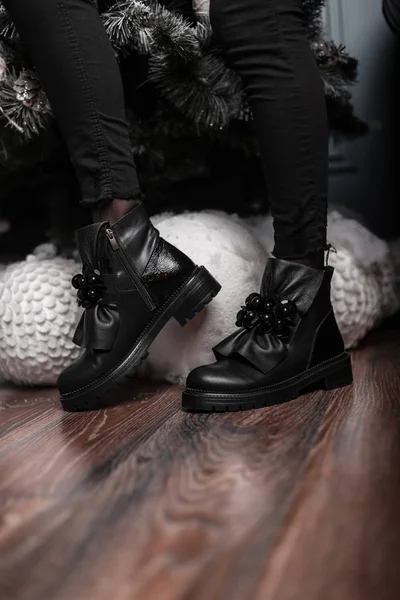 Bottes d'hiver jeunes femmes noires en cuir à la mode. Élégante jeune femme en jeans dans des chaussures à la mode se trouve dans une pièce près de belles boules de Noël blanches. Nouvelle collection de chaussures saisonnières . — Photo
