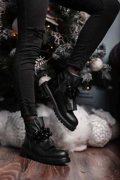 Trendy young woman in vintage jeans in fashionable leather black shoes poses near a Christmas tree in the room. Closeup of female legs in stylish boots. Winter fashion youth footwear. — Stock Photo, Image