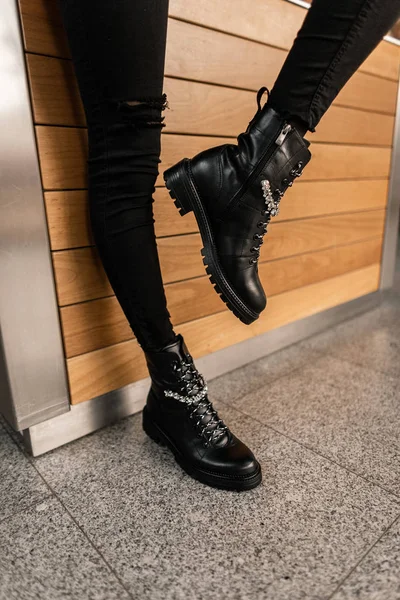 Jeune femme à la mode en jean vintage noir avec des bottes élégantes en cuir avec des strass en dentelle posant près d'un mur en bois dans le studio. Gros plan des jambes féminines dans des chaussures saisonnières tendance. Mode . — Photo