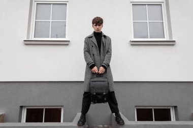 Şık, ekose ceketli, şık bir gencin model modeli. Şehirde beyaz bir duvarın yanında siyah deri bir sırt çantasıyla modaya uygun botları var. Modern yakışıklı adam. Sonbahar-kış tarzı. Erkek giyim.
