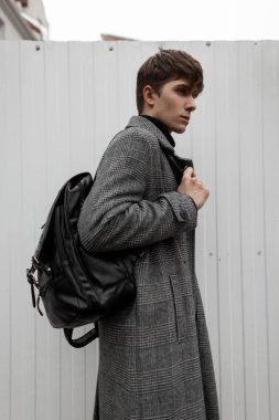 Gri ekose ceketli, şık saç stili, deri sırt çantalı Avrupalı genç adam şehirde metal beyaz bir duvarın yanında duruyor. Çekici adam bir manken. Modaya uygun erkek giyimi.
