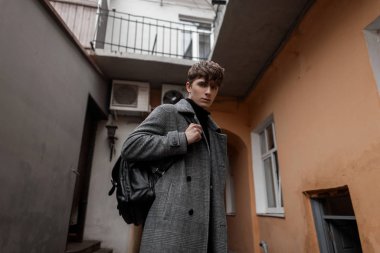 Modern moda genç adam modeli şehrin her yerinde gri kareli montlu deri siyah bir sırt çantasıyla dolaşıyor. Sokakta şık bir saç stili olan yakışıklı bir adam. Avrupa tarzı gençlik.