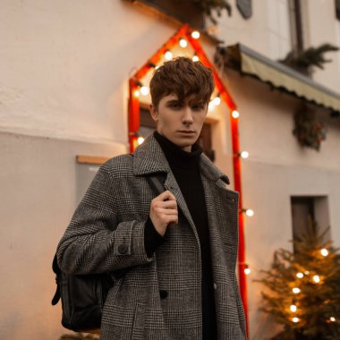 Son moda ekose ceketli yakışıklı, deri sırt çantalı genç adam çelenk ve Noel ağacıyla süslenmiş bir binanın yakınındaki bir sokakta duruyor. Yakışıklı adam dışarıda dolaşmaktan zevk alıyor..