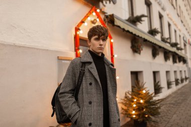 Şık kış kıyafetleri içinde şık bir genç adam şehrin dışında, klasik ışıklar ve Noel ağacıyla süslenmiş bir binanın yanında duruyor. Şık giyinmiş yakışıklı sokakta poz veriyor.