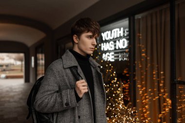 Omzunda şık bir sırt çantası olan gri kareli sezonluk modaya uygun montu olan son moda genç adam, sokakta parlak ışıklı bir dükkan penceresinin yanında duruyor. Şehirdeki adam.