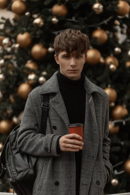 Eski bir Noel ağacının arka planında elinde bir fincan kahveyle şık kışlık giysiler içinde genç ve yakışıklı bir adam duruyor. Şehirdeki zarif dış giysili çekici bir adam..