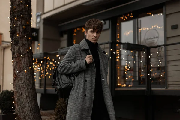 Sexy stijlvolle jongeman in modieuze winterkleding met een trendy rugzak staat in de stad in de buurt van een café ingericht met nieuwjaar vintage lichten. Modern mooi mannenmodel op straat. Jongerenmode. — Stockfoto