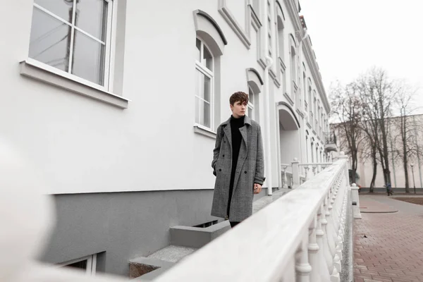 Stijlvolle aantrekkelijke jongeman in een modieuze grijze jas met een trendy kapsel wandelingen op de stad in de buurt van een wit gebouw. Moderne man fashion model.Nieuwe collectie van mannen herfst elegante buitenkleding — Stockfoto