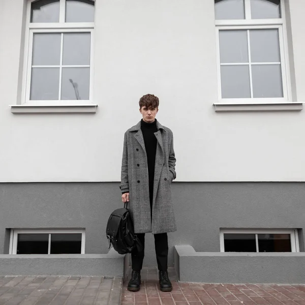 Ελκυστικός νεαρός άνδρας σε ένα μακρύ καρό μοντέρνο παλτό σε μαύρο κομψό μπότες με ένα δροσερό δερμάτινο σακίδιο στέκεται στο δρόμο κοντά σε ένα λευκό κτίριο. Όμορφος Ευρωπαίος έξω. Ανδρικά ενδύματα. — Φωτογραφία Αρχείου