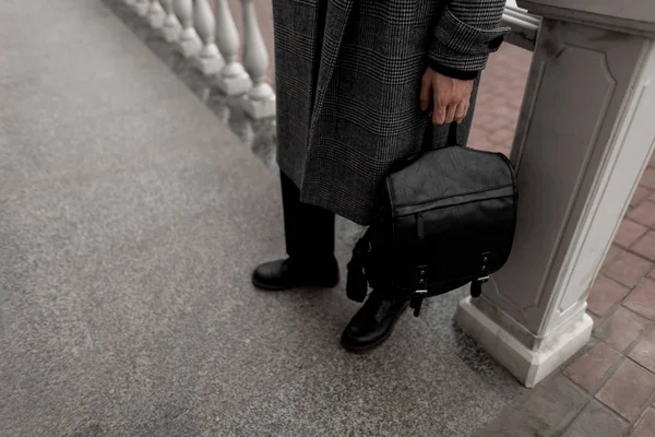 Primer plano de las piernas del hombre elegante en botas brutales con estilo en un largo elegante abrigo a cuadros de moda al aire libre. Urbano chico está de pie en la calle con una mochila de cuero vintage negro. Moda casual juvenil . — Foto de Stock