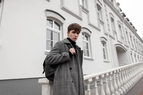 Elegant handsome young man in a fashionable plaid coat with a stylish leather backpack with a trendy hairstyle stands on the street near a building on an autumn day. Modern men's seasonal outerwear. — 스톡 사진