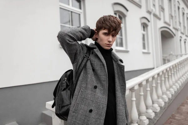 Chico sexy en ropa interior otoño-invierno estira el cabello con estilo. Modelo de hombre atractivo joven europeo en un abrigo a cuadros gris con una mochila de cuero de moda posa al aire libre cerca de un edificio blanco vintage . — Foto de Stock