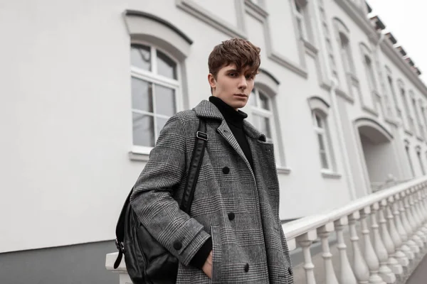 Portrait of a serious handsome young man in elegant checkered gray coat with leather black vintage backpack poses outdoors near a white building. Attractive fashion model of a stylish guy. Youth style