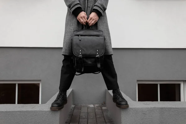 Close-up of a man in a stylish gray plaid coat in elegant pants in winter black boots with a leather backpack in hands near a vintage wall in the city. Trendy new collection of seasonal men's clothing — 스톡 사진