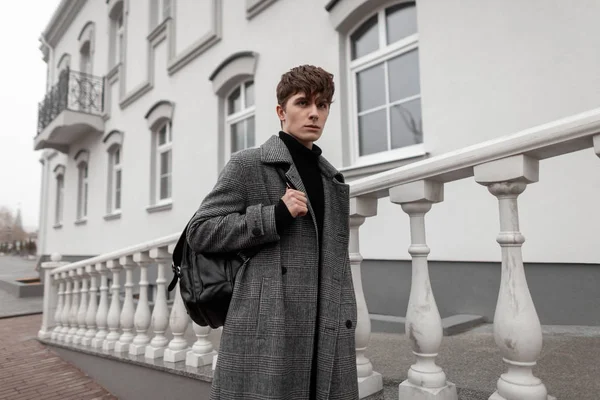 流行のグレーのコートを着たヨーロッパの若者は、ヴィンテージの建物の近くの街にレザーブラックのバックパックを備えたスタイリッシュな髪型をしています。魅力的な男はファッションモデルです。ファッショナブルなメンズウェア — ストック写真