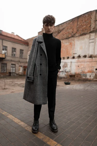 Model of a stylish young man in an autumn fashionable gray check coat in black trendy clothing in vintage leather shoes stands on a street in the city. Attractive modern guy. Elegant men's clothing. — Stock Photo, Image