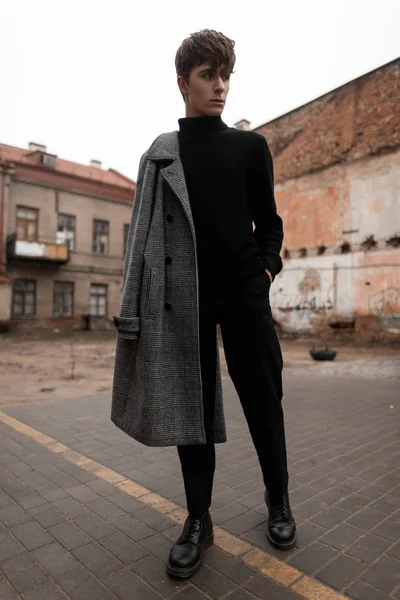 Young European man model with a fashionable hairstyle in a stylish gray plaid coat in jeans in a black vintage sweater stands outdoors on the street among the buildings. Handsome trendy european guy. — Stock Photo, Image