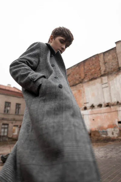 In a fashionable gray plaid long coat a handsome stylish young man with a trendy hairstyle is standing on the street. Attractive guy model in seasonal clothes poses outdoors. New collection menswear. — 스톡 사진