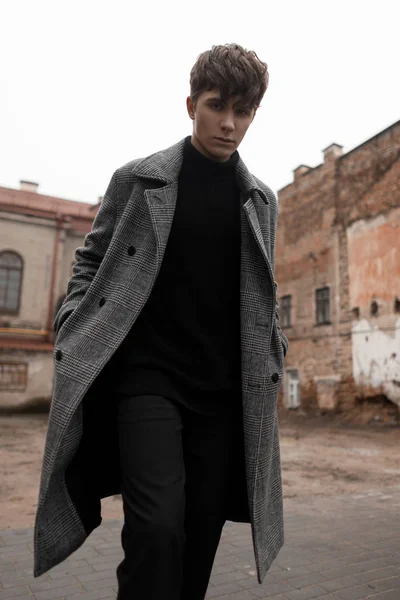 Jonge knappe man met een modieus kapsel in een luxe klassieke geruite jas in een trendy gebreide trui in jeans loopt door de straat in de oude stad. Urban guy in elegante kleding. — Stockfoto