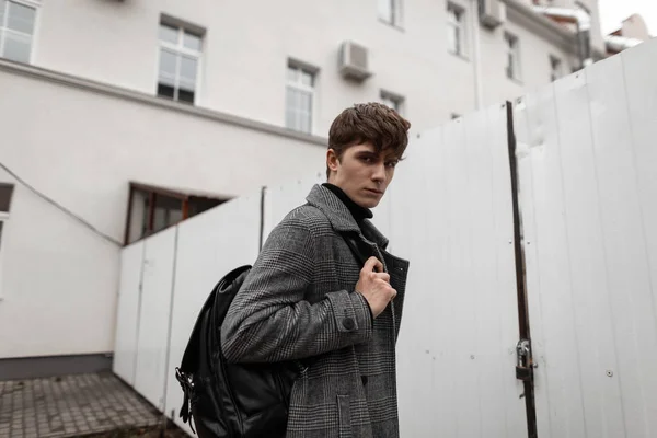 Jonge knappe man model in een modieuze grijze geruite jas met een leren rugzak staat op straat in de buurt van een wit metalen hek. Europese man in stijlvolle buitenkleding wandelingen in de stad. Wintercollectie. — Stockfoto