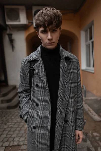 Jeune homme modèle dans un élégant manteau à carreaux gris dans un pull tricoté noir avec une coiffure à la mode posant dans la ville dans la cour d'un bâtiment vintage. Charmant mec en vêtements élégants à l'extérieur. Style . — Photo