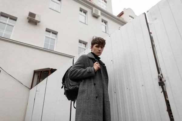 In een modieuze geruite jas met een stijlvolle leren rugzak loopt een knappe jongeman model langs een straat in de buurt van een wit metalen hek. Trendy moderne jongen in de jeugd herfst-winter kleding buiten. — Stockfoto