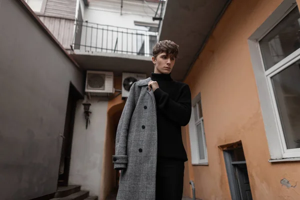 Trendy όμορφος νεαρός άνδρας σε ένα μοντέρνο καρό γκρι παλτό σε μαύρο κομψό πουλόβερ στέκεται σε εξωτερικούς χώρους κοντά στο vintage τοίχους. Ελκυστικός μοντέρνος τύπος ποζάρει με κομψά ρούχα. Νεανική μοντέρνα εμφάνιση. — Φωτογραφία Αρχείου
