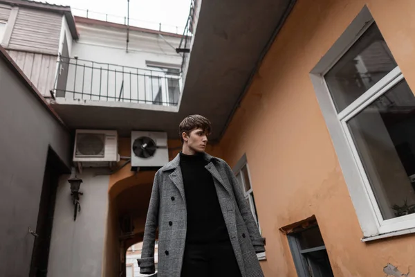 Joven guapo con un peinado elegante en un clásico abrigo a cuadros gris de moda en un suéter de punto negro se encuentra en el patio de un edificio antiguo. Modelo de moda atractivo chico elegante . — Foto de Stock