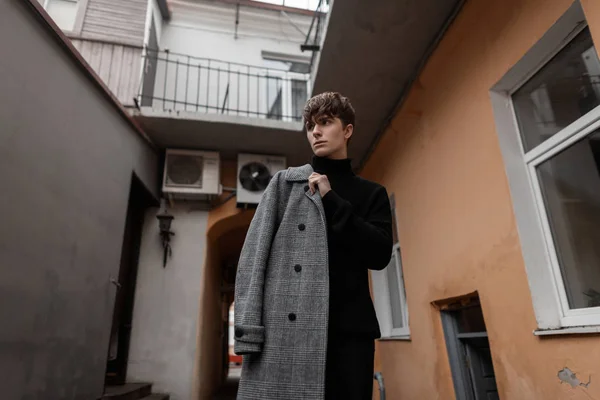 Atractivo joven urbano con ropa negra con estilo en un clásico de moda abrigo gris a cuadros posando al aire libre cerca de un edificio vintage en un cálido día de otoño. Guapo modelo chico de moda al aire libre. Estilo . — Foto de Stock