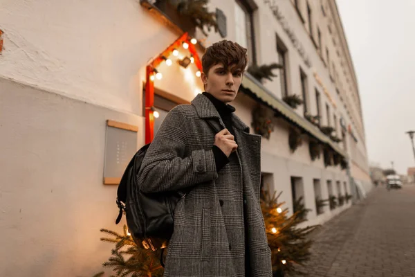 Módní mladý muž se stylovým účesem v módním elegantním zimním kabátě stojí na ulici u zdi s vánoční výzdobou. Módní evropský model si užívá procházky v zimním dni. — Stock fotografie