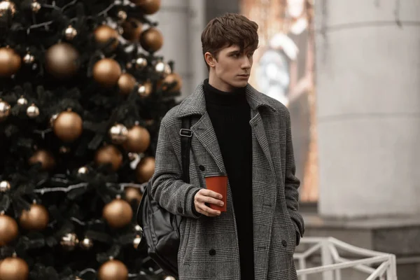 Hipster atractivo hombre en un elegante abrigo a cuadros con un peinado elegante se levanta y sostiene en la mano una taza con un té caliente cerca del árbol de Año Nuevo festivo con juguetes vintage. Chico guapo modelo en la ciudad — Foto de Stock