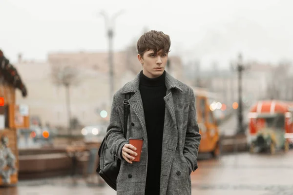 Modelo de un joven de moda en un elegante abrigo a cuadros juveniles con un peinado de moda se encuentra en la plaza de la ciudad con una bebida caliente y sabrosa en las manos. chico urbano camina al aire libre y bebe té dulce . — Foto de Stock